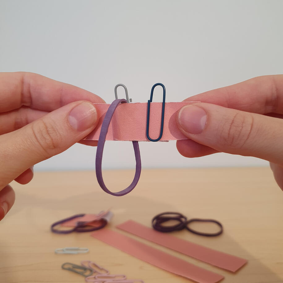 Hands linking paperclips and a rubber band on a folded paper strip, a topology magic trick showing how loops link when pulled taut