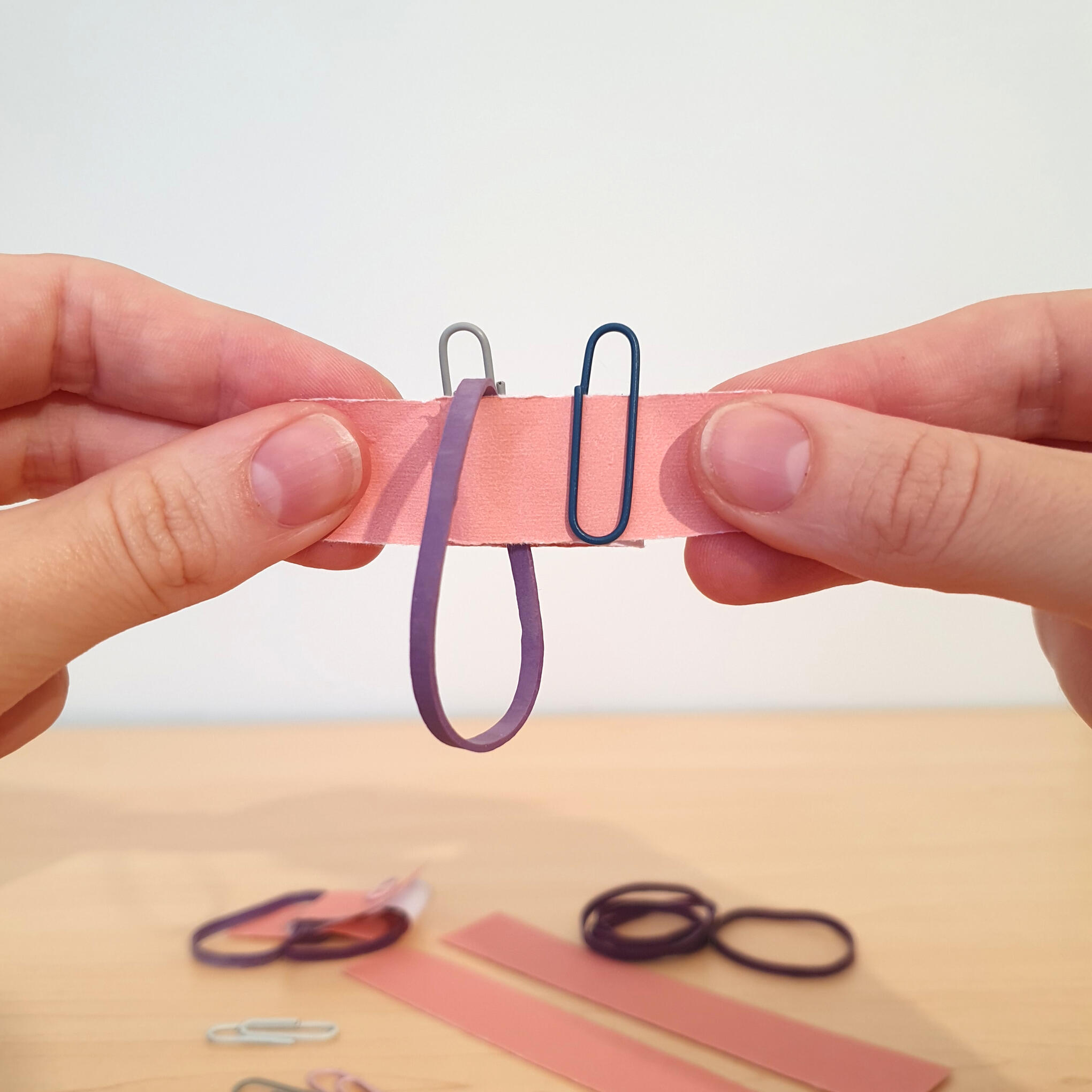 Hands linking paperclips and a rubber band on a folded paper strip, a topology magic trick showing how loops link when pulled taut